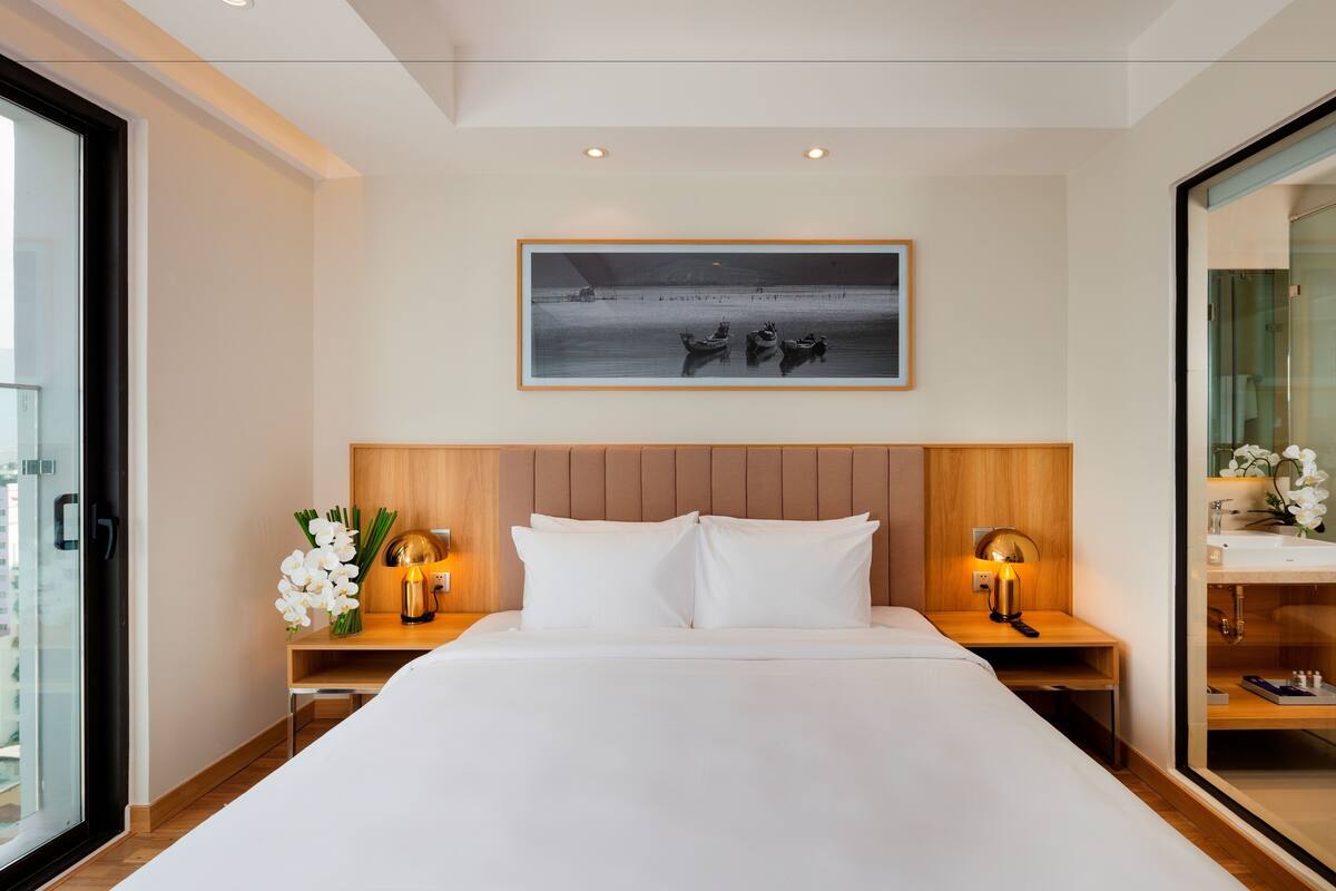 Habitación de hotel con una cama grande, dos mesitas de noche con lámparas, una obra de arte enmarcada sobre la cama y vistas a través de un ventanal.