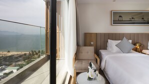 Ein Hotelzimmer mit großem Fenster und Blick auf einen Strand, ein Bett, ein Sessel und ein kleiner runder Tisch mit Blumen.