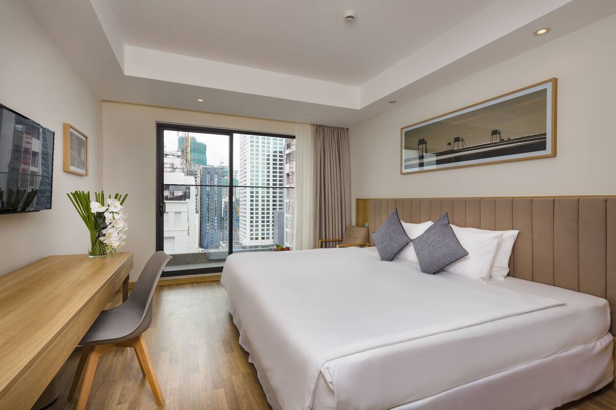 Habitación de hotel moderna con una cama grande, un escritorio, una silla y vistas de la ciudad a través de un ventanal amplio.