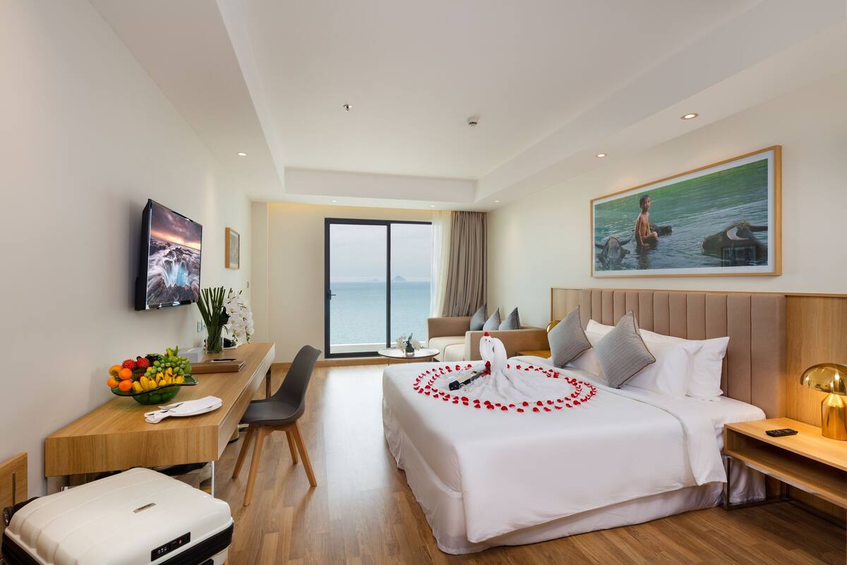 Habitación de hotel con cama, escritorio con frutero, una silla, maleta, televisor y un cuadro de una persona con elefantes.