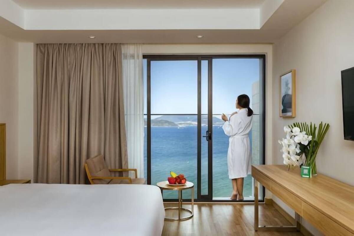 Habitación de hotel con cama, una silla, una mesita con fruta, un florero y vistas al mar a través de una puerta corredera.