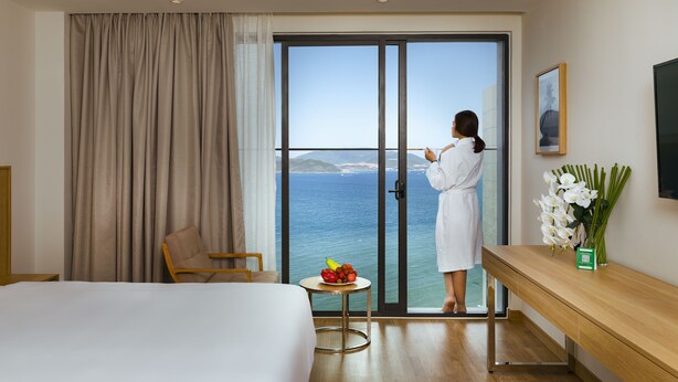 Ein Hotelzimmer mit Bett, Sessel, kleinem Tisch mit Obst, Blumenvase und Meerblick durch eine Schiebetür.