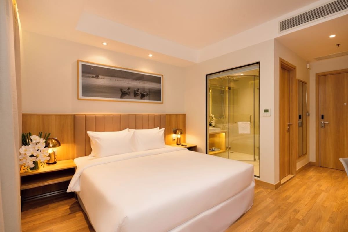 A hotel room with a large bed, a bedside table with a lamp, a framed picture on the wall, and a glass-enclosed shower area.