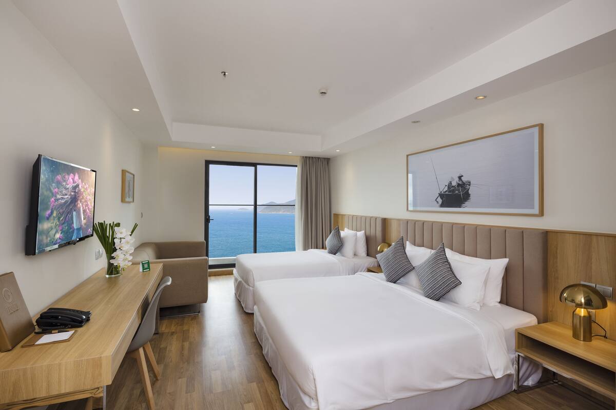 Habitación de hotel con dos camas, un escritorio, una silla, un televisor y un ventanal con vistas al mar.