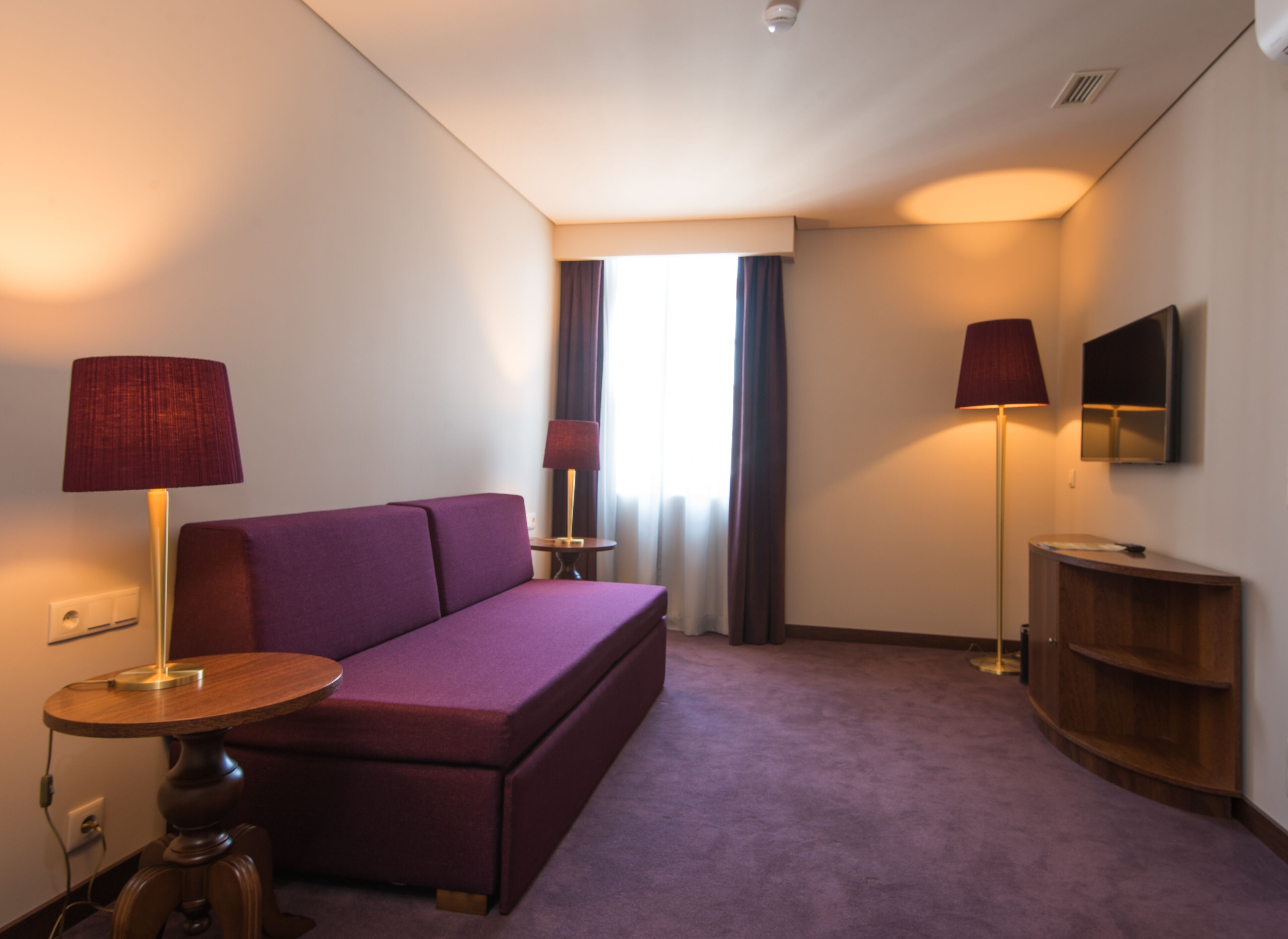 A hotel room with a purple sofa, two lamps, a TV, and a wooden cabinet.