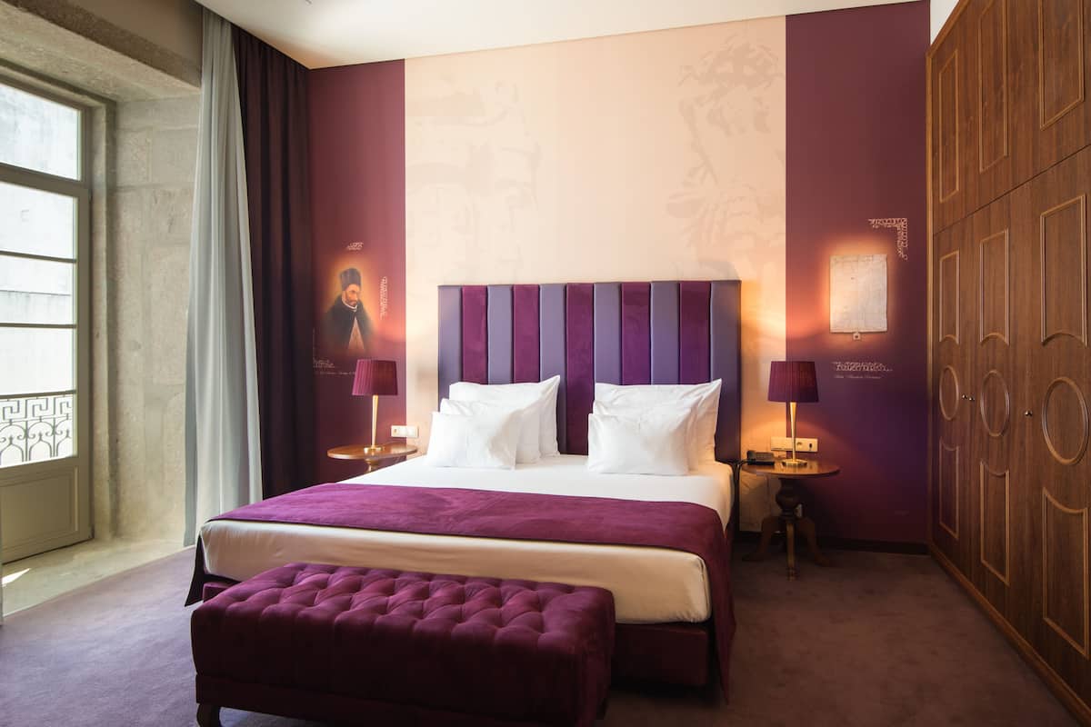 A hotel room with a large bed, a bedside table, a lamp, a wardrobe, and a window with curtains.