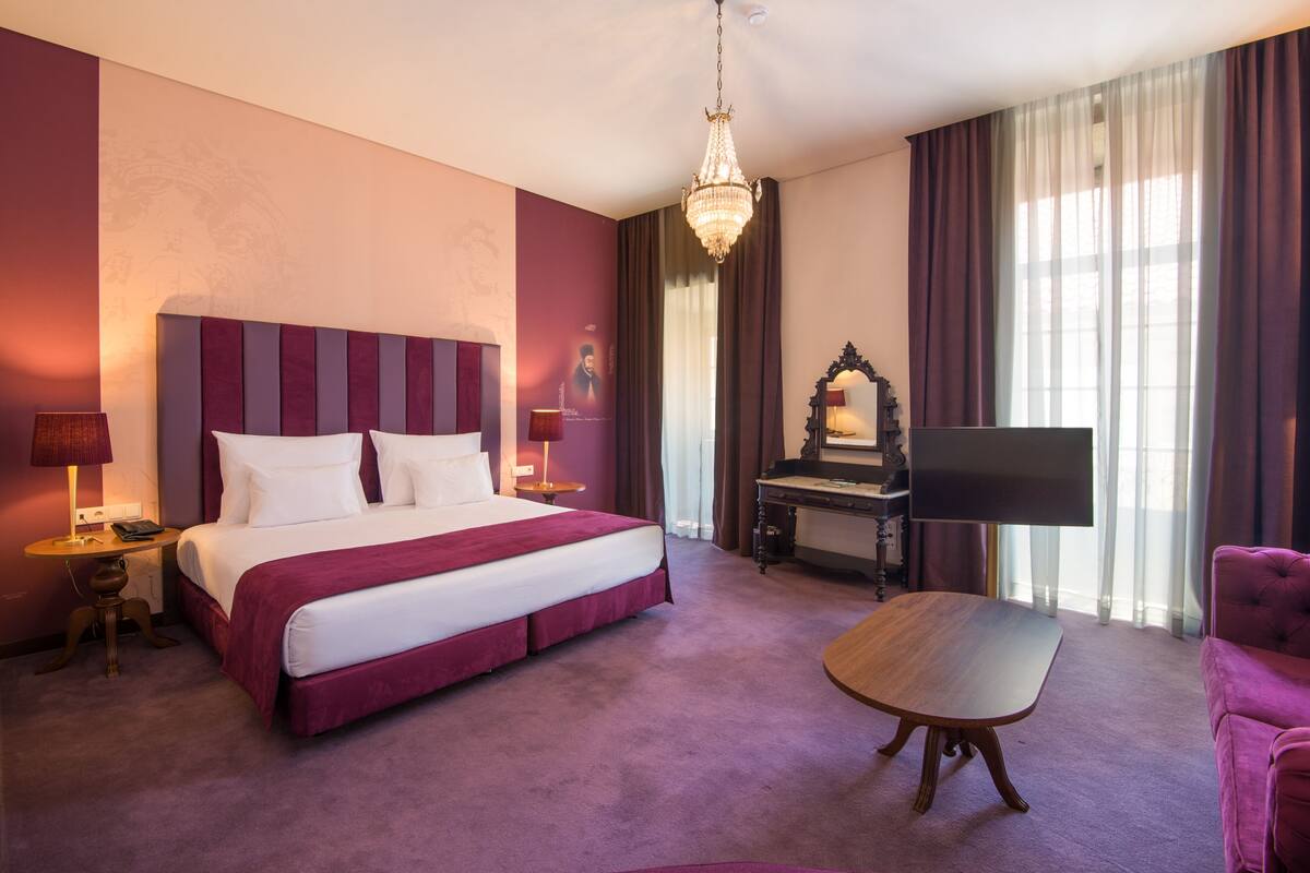 A hotel room with a large bed, a flat-screen TV, a wooden coffee table, and a pink armchair.