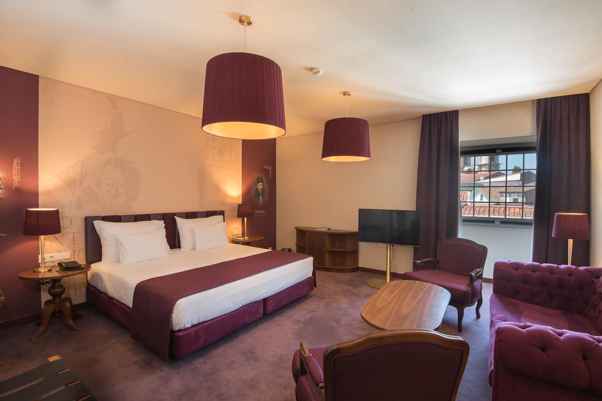 A hotel room with a large bed, a desk, a chair, a TV, and a window with curtains.