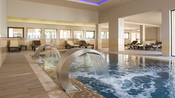 Une piscine intérieure dotée d’un élément décoratif aquatique moderne, de chaises longues et de grandes fenêtres.
