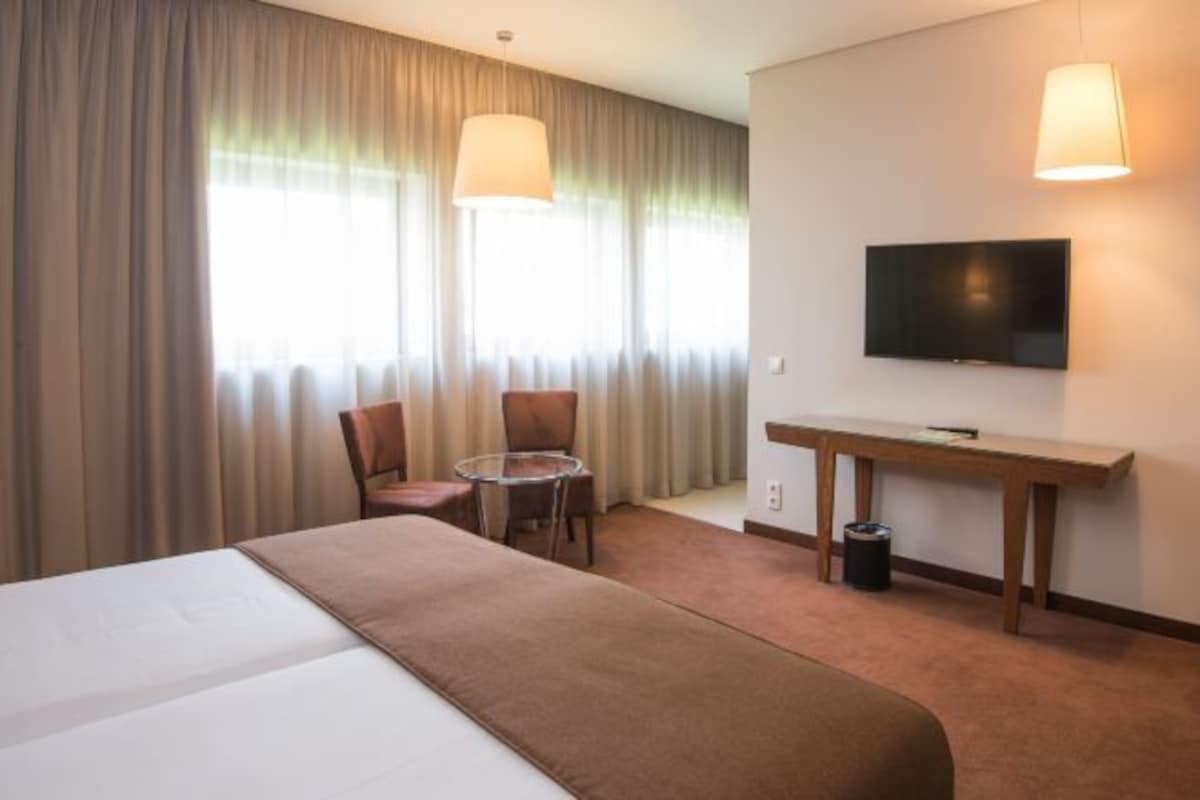 Una habitación de hotel con cama, escritorio, silla, televisor y ventana con cortinas.