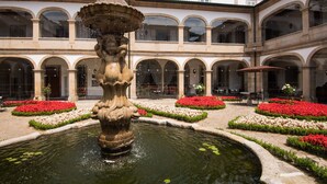 Une cour intérieure avec une fontaine centrale, entourée de parterres de fleurs et d’un bâtiment doté de fenêtres en arc de cercle.