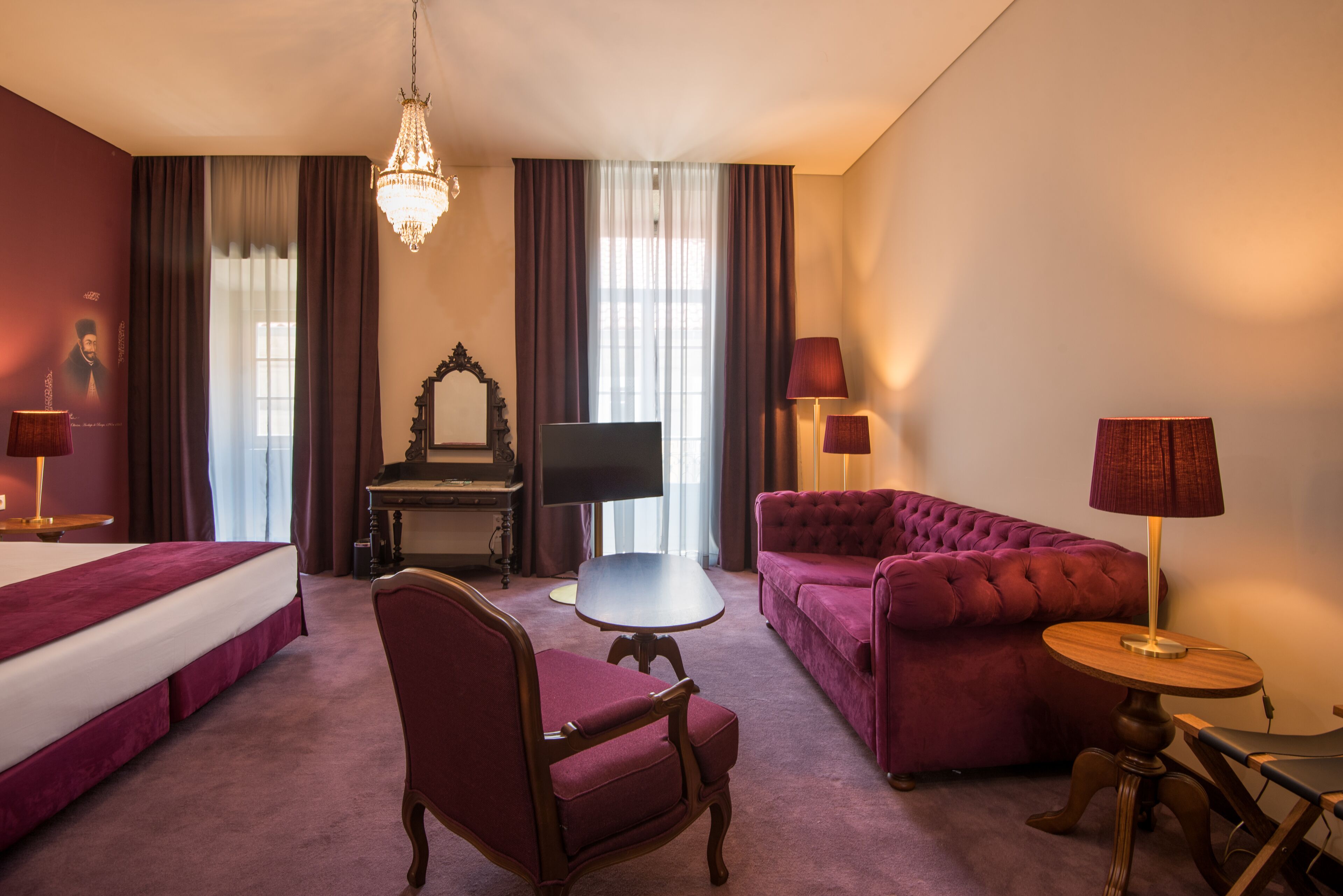 Una camera d'albergo con un letto, un divano, una poltrona, un tavolino, una TV, un lampadario e tende.