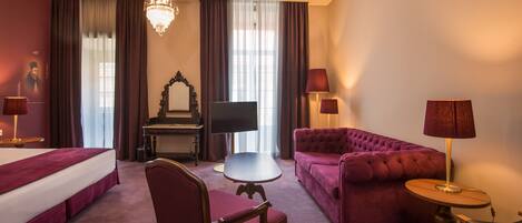 A hotel room with a bed, a sofa, a chair, a small table, a TV, a chandelier, and curtains.