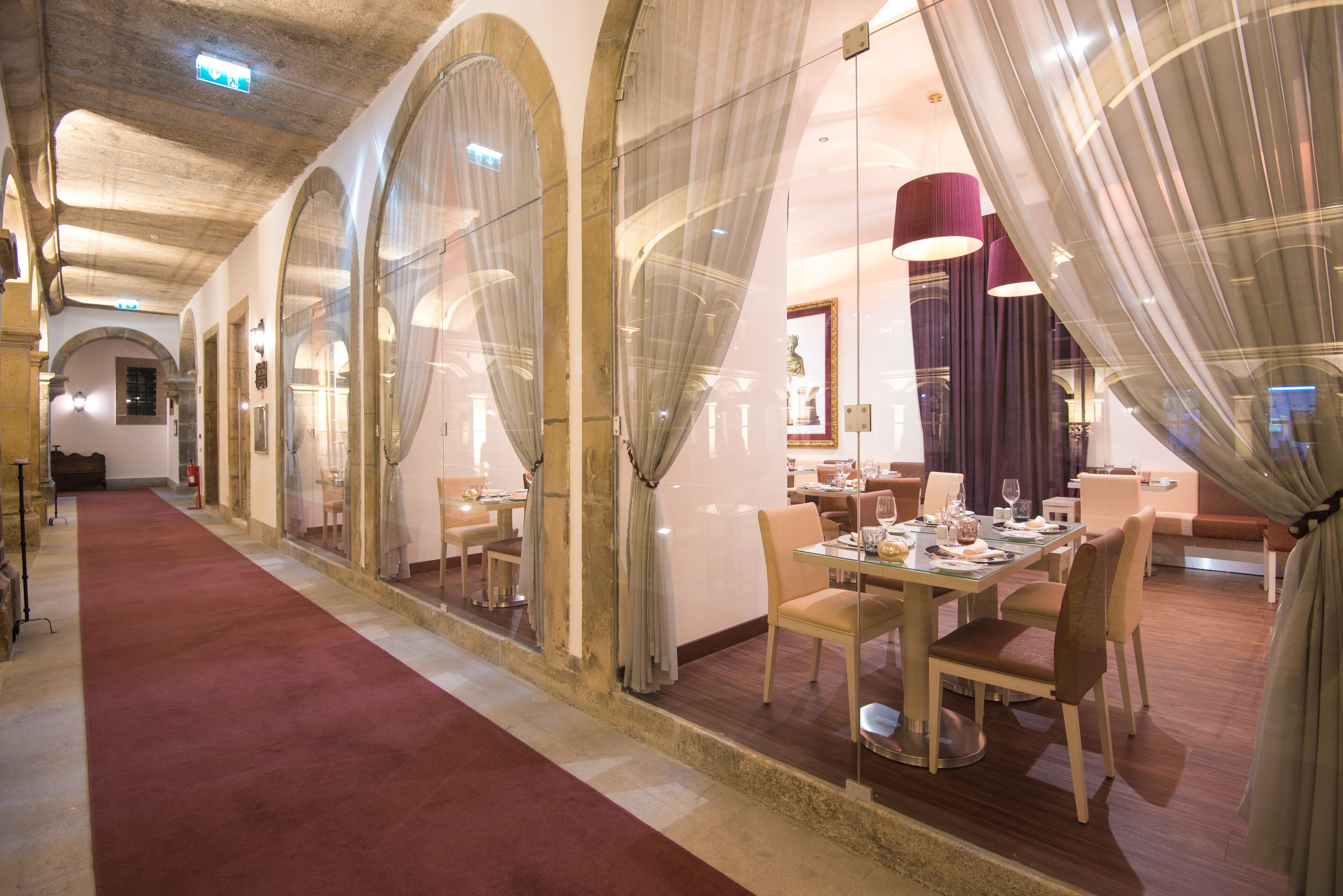 A hotel corridor with arched glass doors, a red carpet, and a dining area with tables and chairs.