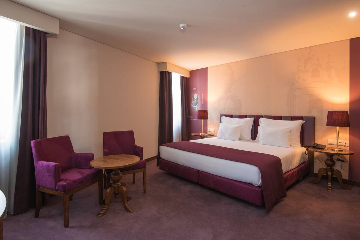 A hotel room with a large bed, two chairs, a small table, and a nightstand with a lamp.
