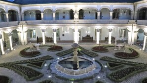 Une cour intérieure avec une fontaine centrale, entourée de parterres de jardin aux formes géométriques et d’un bâtiment à plusieurs niveaux doté de fenêtres en arc de cercle.