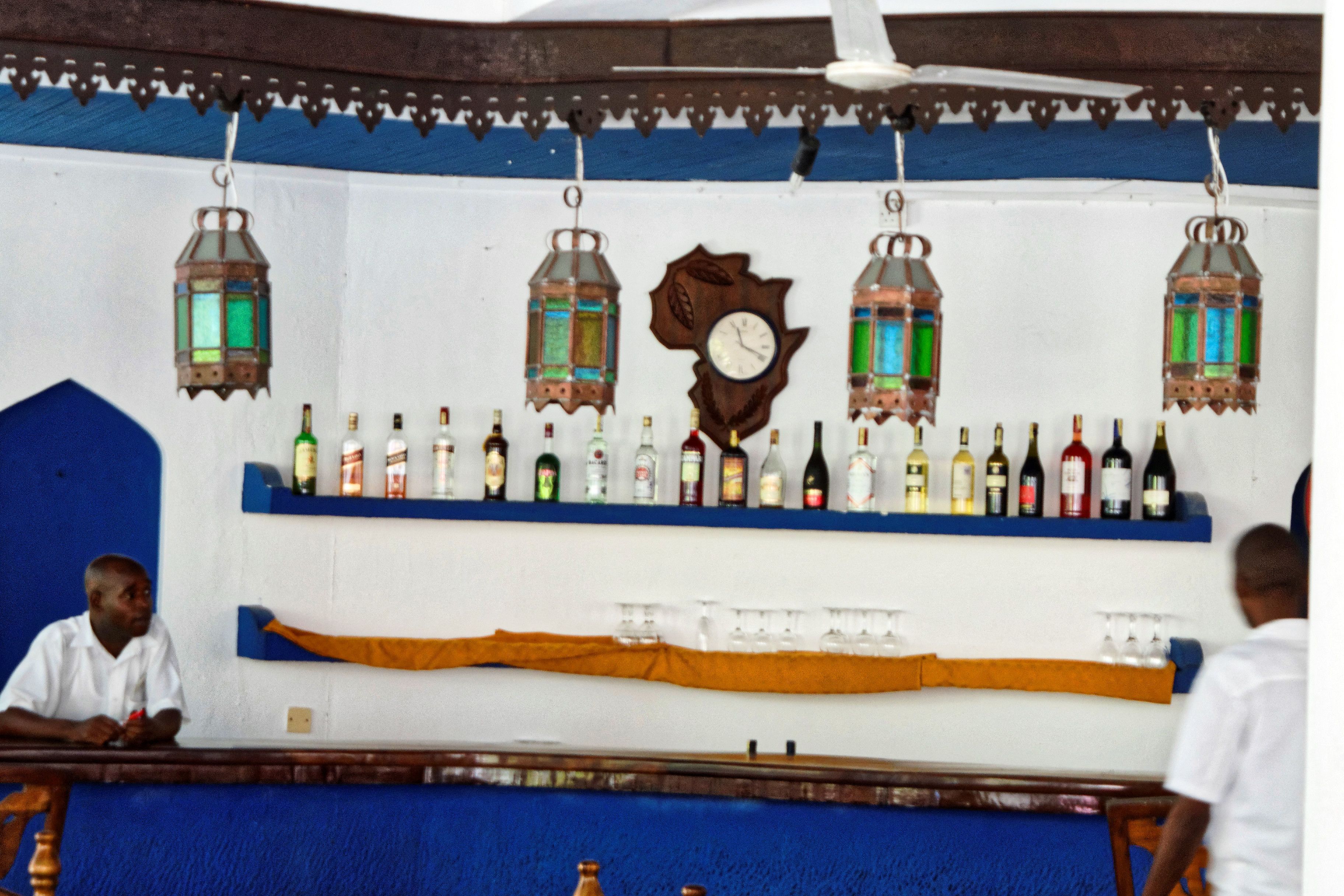 A bar area with a clock, hanging lanterns, and shelves of bottles.