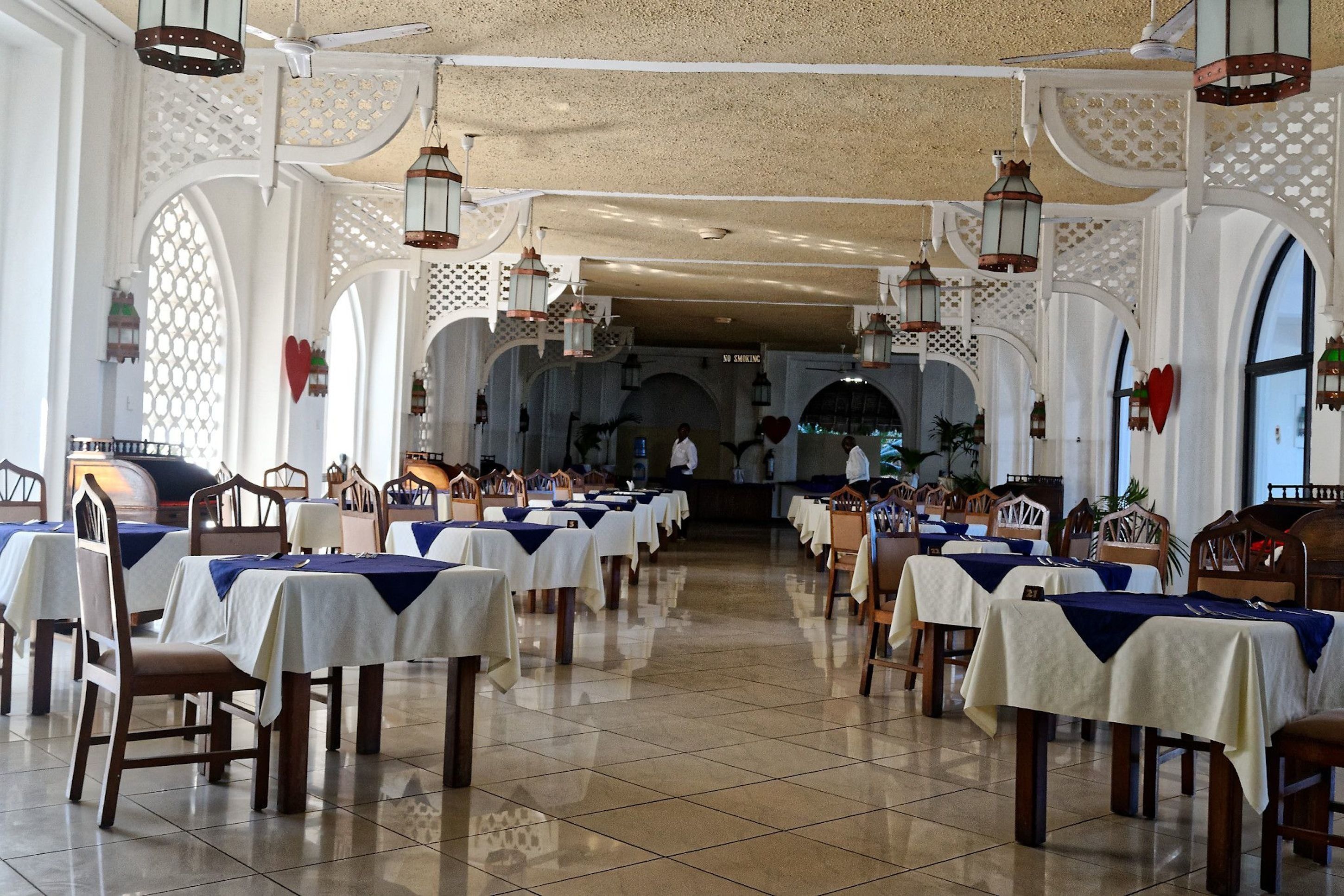 Uma ampla área de refeições com mesas preparadas para o serviço, paredes brancas e lanternas decorativas.