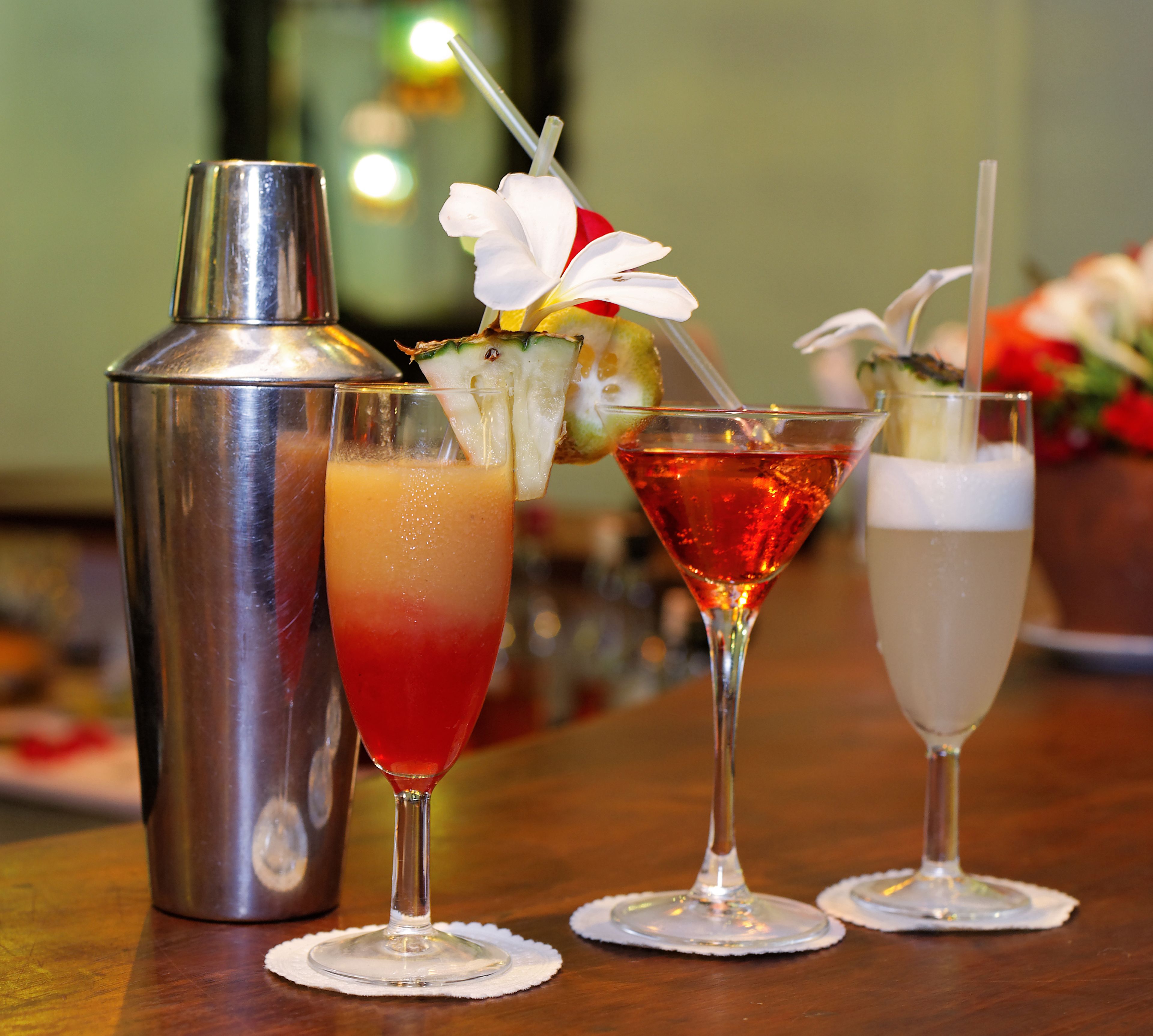 Trois cocktails sont posés sur le comptoir d’un bar, avec un shaker et une bouteille en arrière-plan.