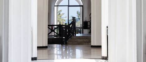 A spacious hallway with white walls and a tiled floor, leading to a room with a large window and a view of palm trees.