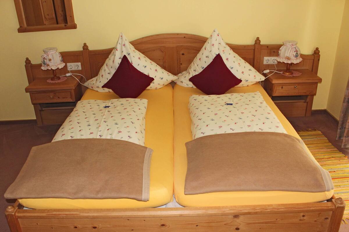 Cama doble con almohadas de estampado floral y una manta marrón, cabecero de madera, mesitas de noche con lámparas y una alfombra estampada.