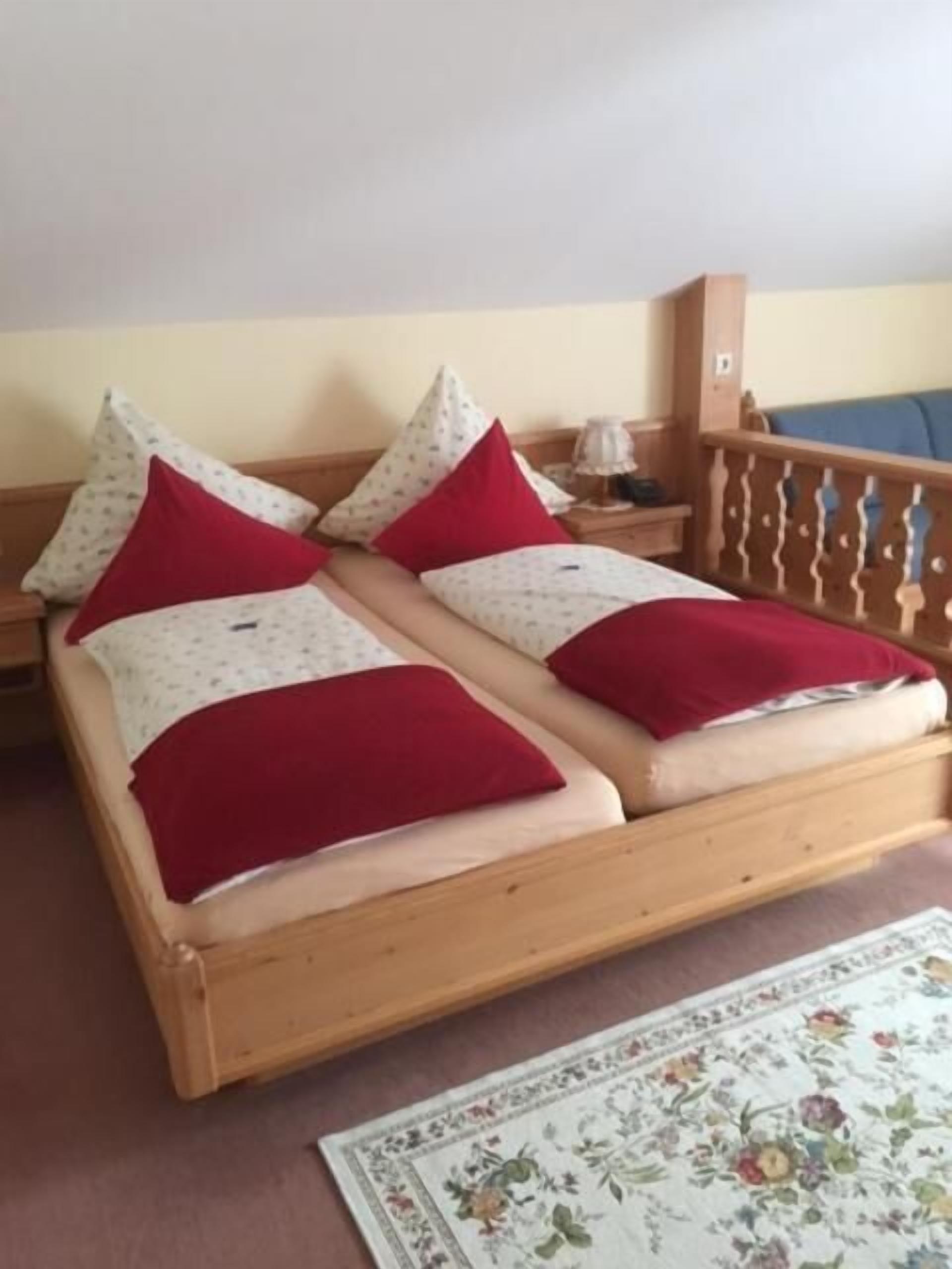 Una cama doble con ropa de cama roja y blanca, un cabecero de madera, una mesita de noche con lámpara y una alfombra estampada con motivos florales.