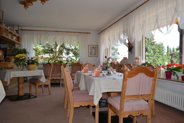 A dining room with wooden chairs and tables, white curtains, and potted plants. - Hotel Adlerschanze (Schoenwald)