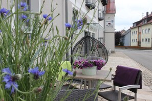 Exterior de un hotel con una mesa, sillas, un arreglo floral y un cartel que indica un área de estacionamiento.