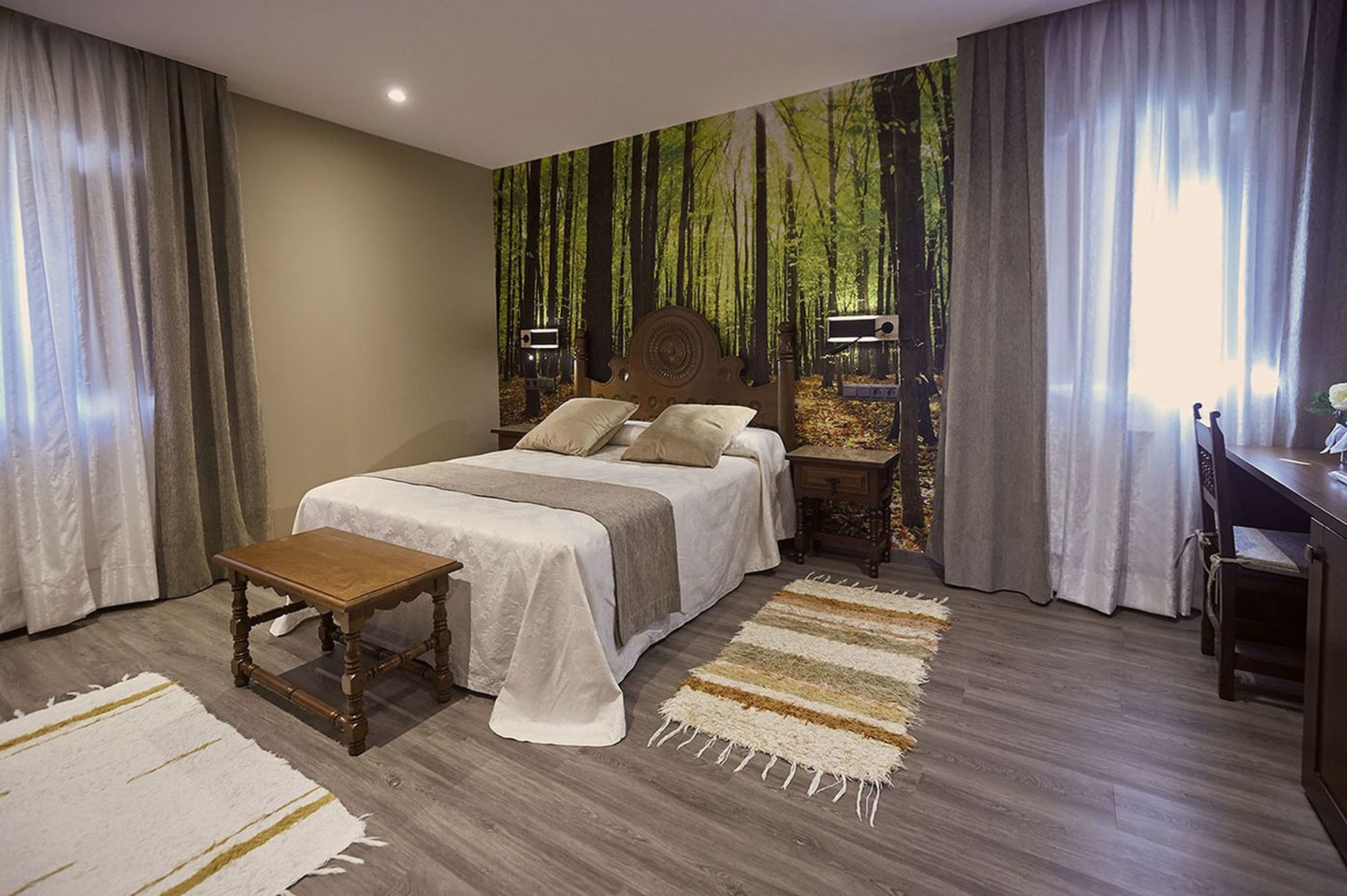 A bedroom with a bed, bedside tables, a nightstand, a wooden bench, and a large forest-themed mural on the wall.