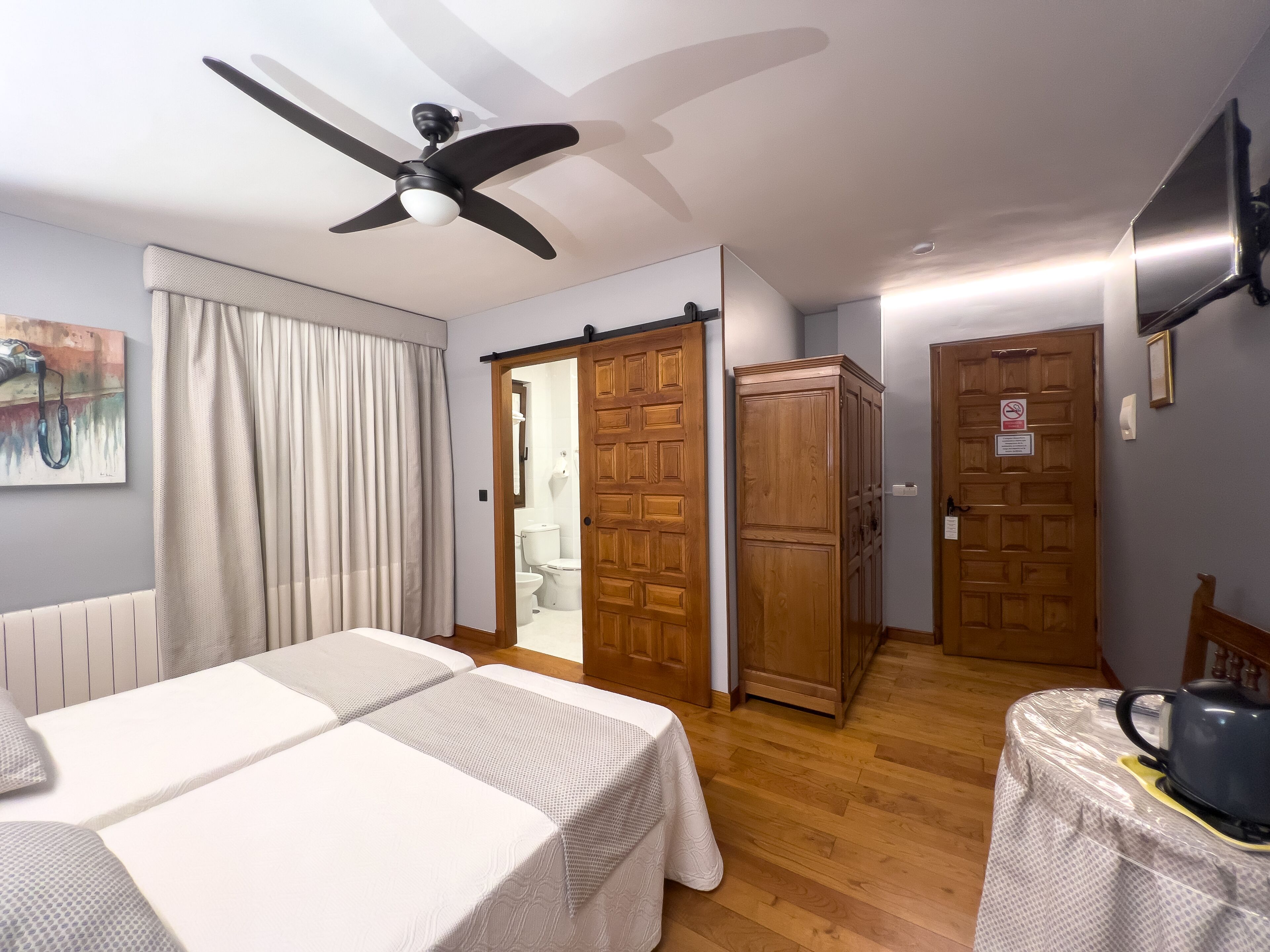A room with two beds, a ceiling fan, wooden doors, and a painting on the wall.