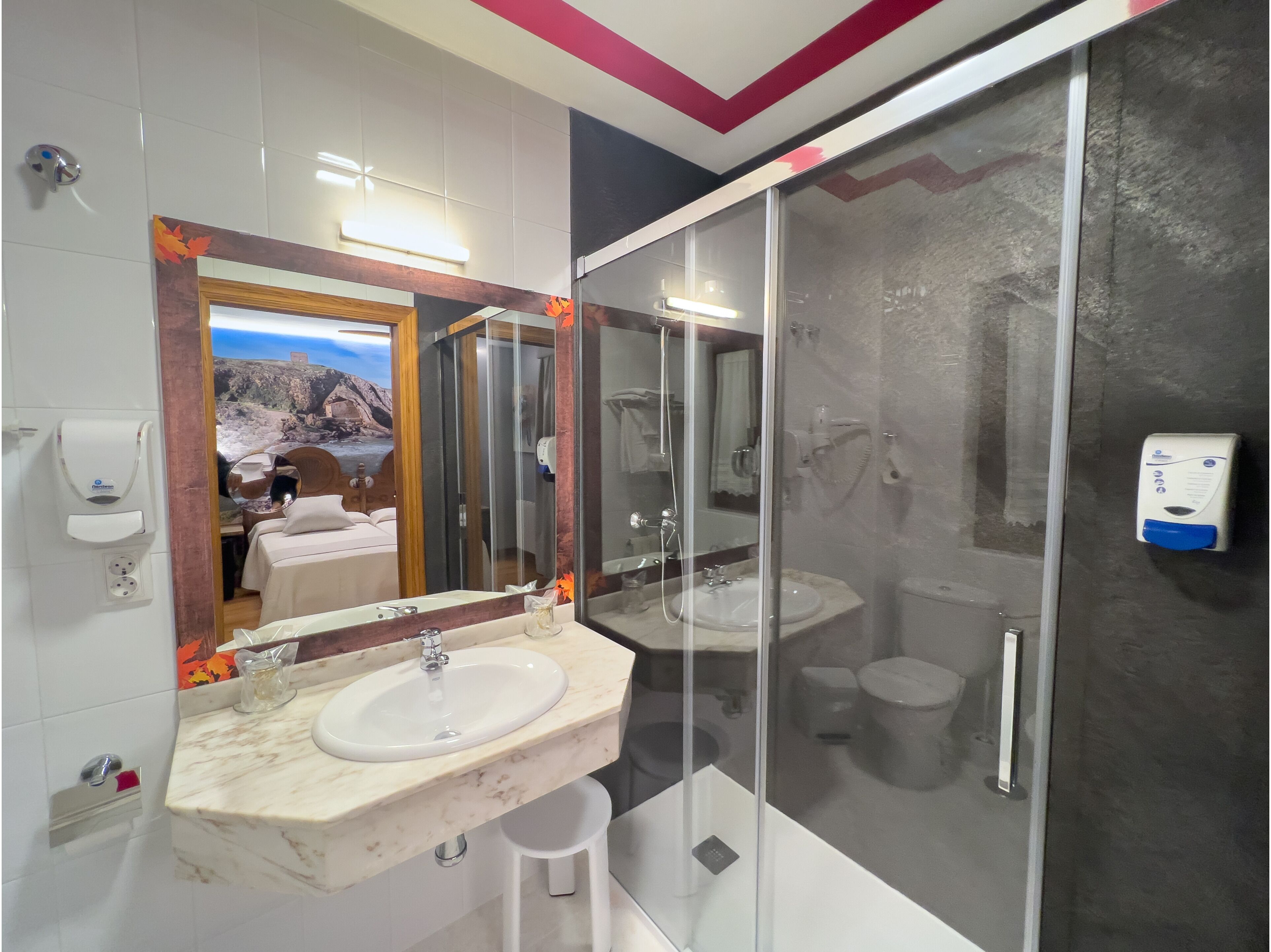 Un baño moderno con lavamanos de mármol, ducha con mampara de vidrio y un espejo grande que refleja un dormitorio con vistas panorámicas.