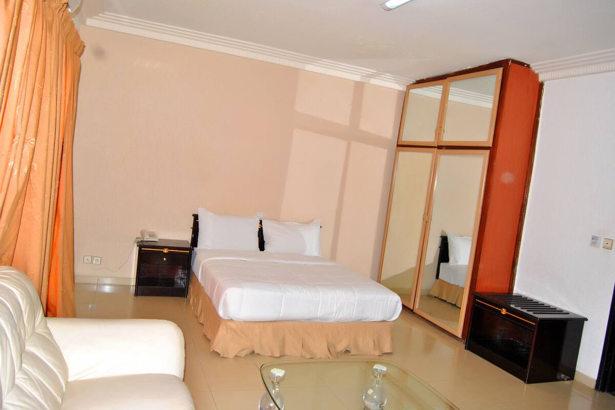 A hotel room with a bed, a nightstand, a mirror, a glass coffee table, and a white sofa.