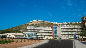 A modern hotel with a central glass facade, multiple balconies, and a view of a hillside with other buildings.