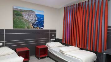 Une chambre d’hôtel avec un lit, deux tables de chevet, un tableau représentant une falaise et l’océan, et des rideaux rouges.