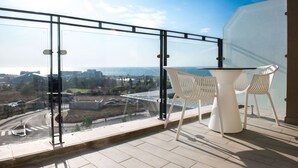 A balcony with a glass railing, a white table, and two white chairs, overlooking a cityscape and a body of water. - HI  Haifa Hostel (Haifa)