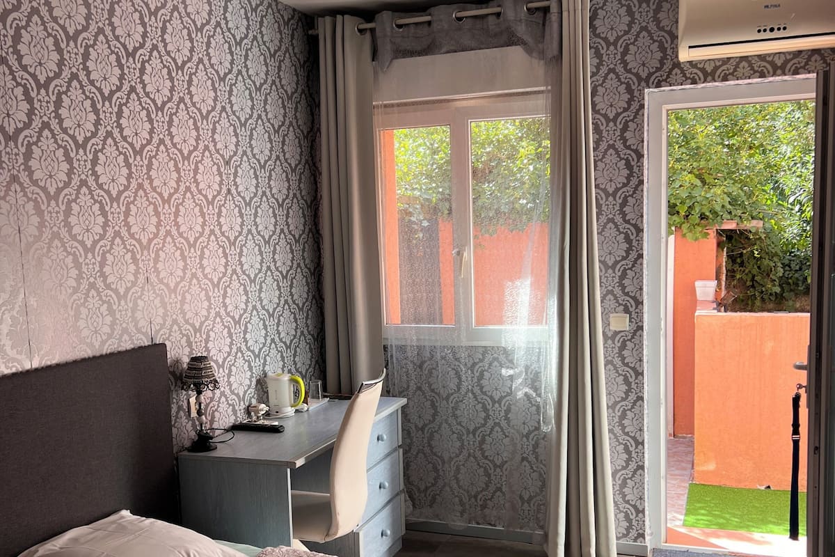 A bedroom with a bed, a desk, and a chair. There is a window with curtains and a view of greenery outside.