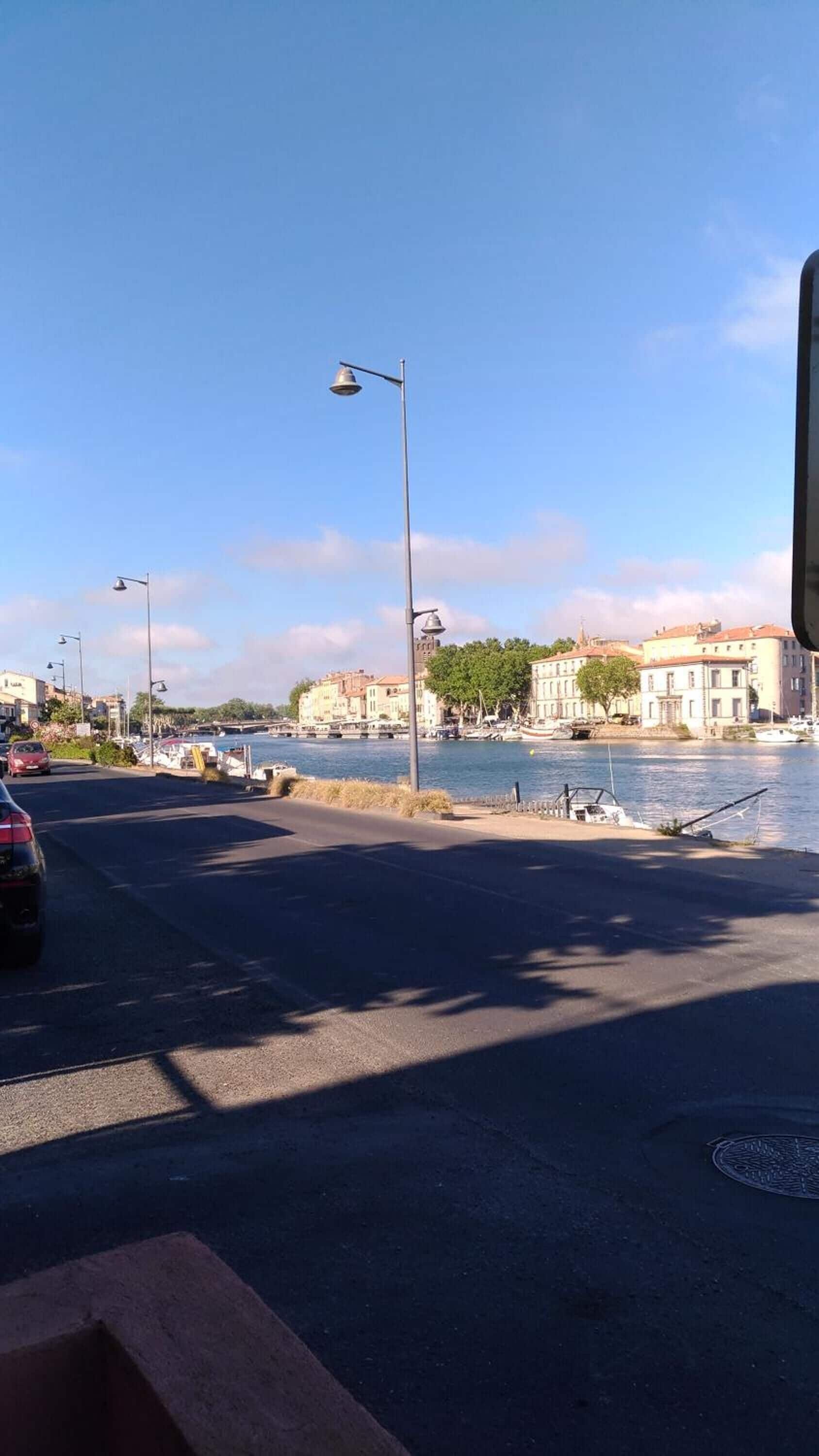 A waterfront street with buildings, a clear sky, and a few parked cars.