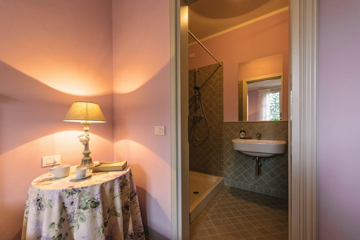Een badkamer met een douche, wastafel en een tafeltje met een lamp en kopjes.