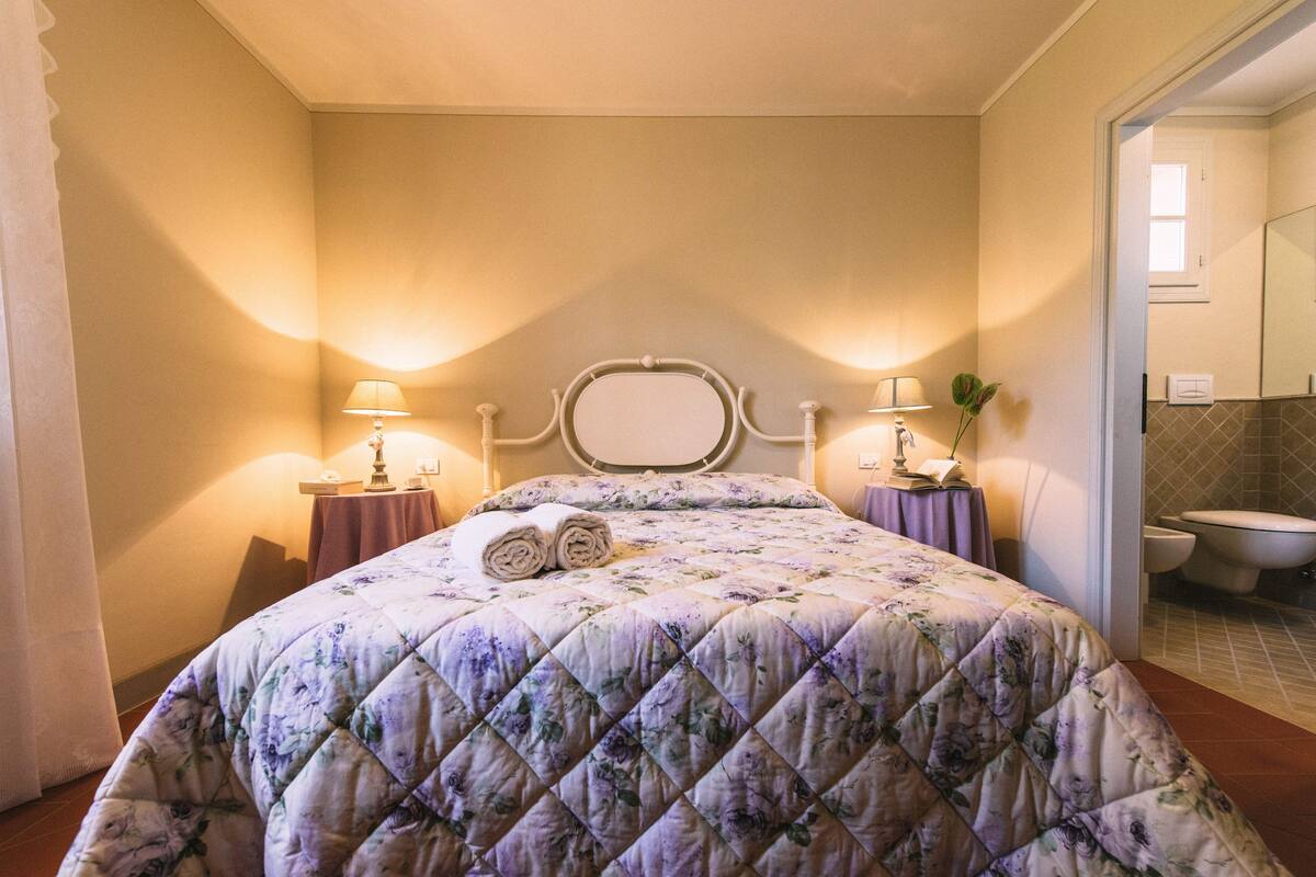 Een net opgemaakt bed met een quilt met bloemenmotief, twee bedlampen en een spiegel boven het bed.