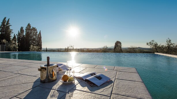 A poolside setting with a bottle, glass, and fruit on a tiled surface, overlooking a clear pool and a scenic landscape.