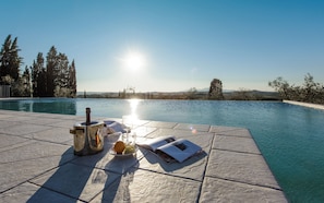Escena junto a la piscina con una botella, un vaso y fruta sobre una superficie de baldosas, con vistas a una piscina cristalina y un paisaje pintoresco.