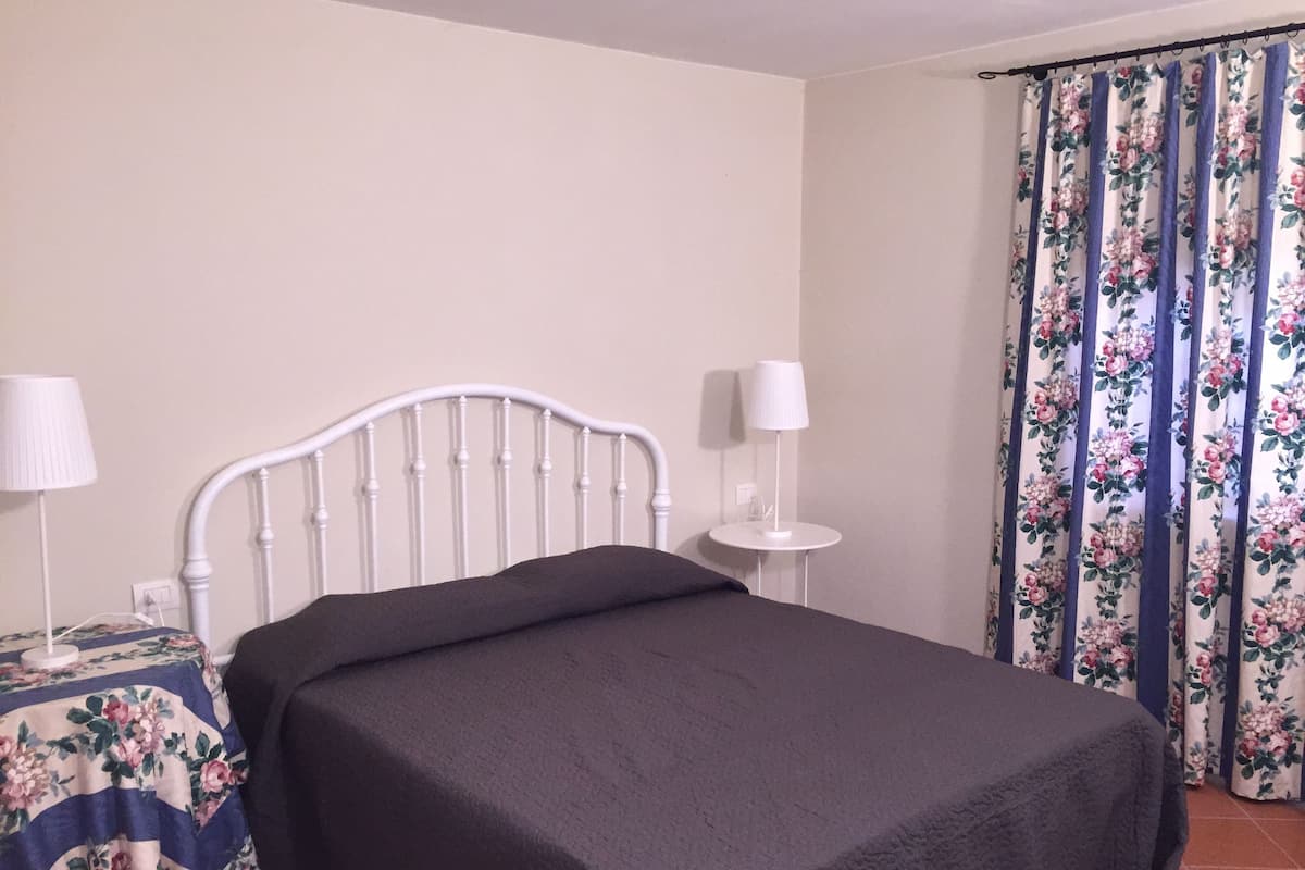 Een bed met een wit metalen frame, een donkergrijze spreidek en een bloemrijk tafelkleed op een nachtkastje.