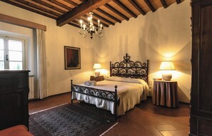 Un dormitorio con cama, dos mesitas de noche con lámparas, una araña y una ventana con cortinas, además de una puerta.