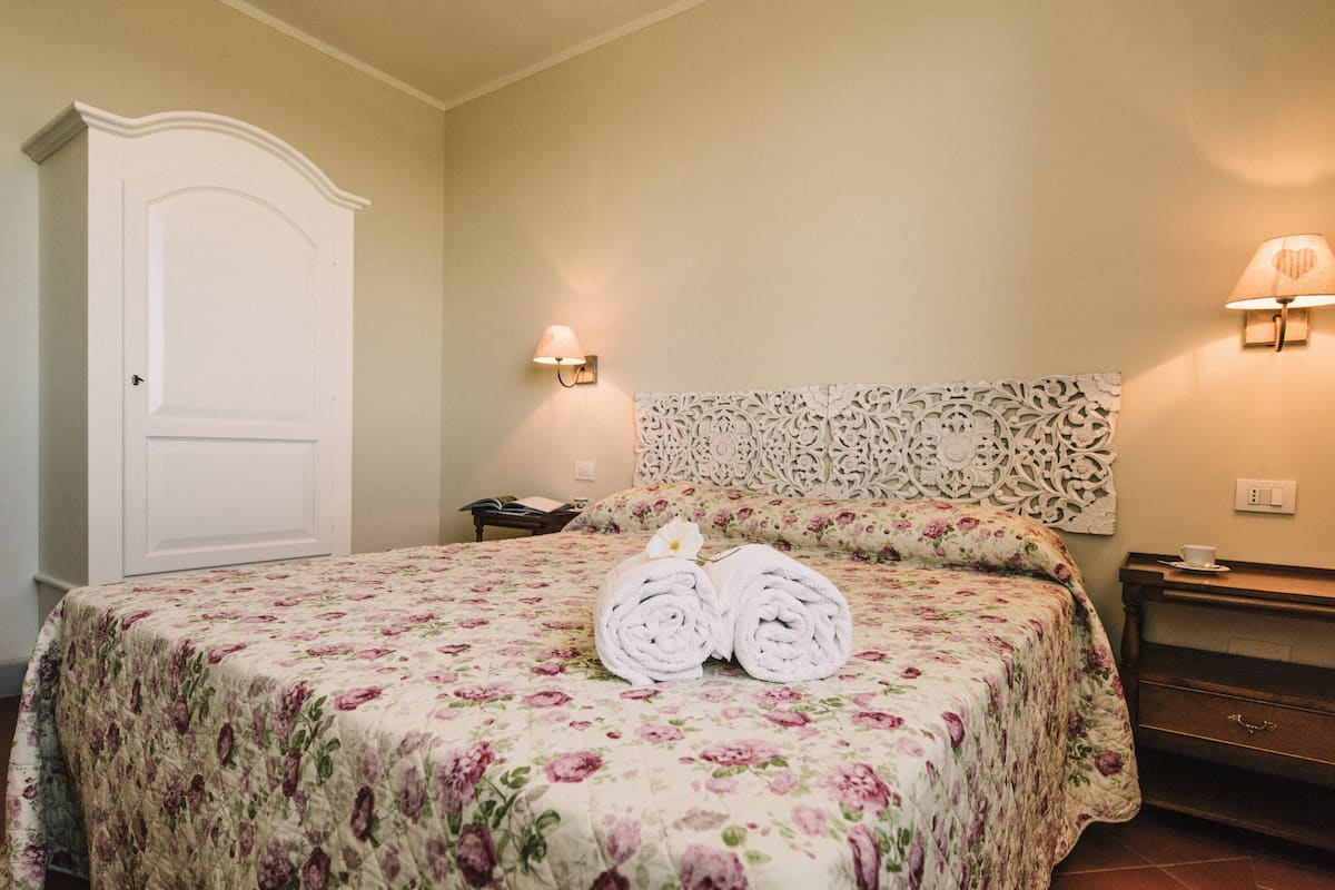 Een hotelkamer met een bloemenbedekker, een decoratieve hoofdbord, twee wandlampen, een nachtkastje met kopje en schotel, en een garderobe.