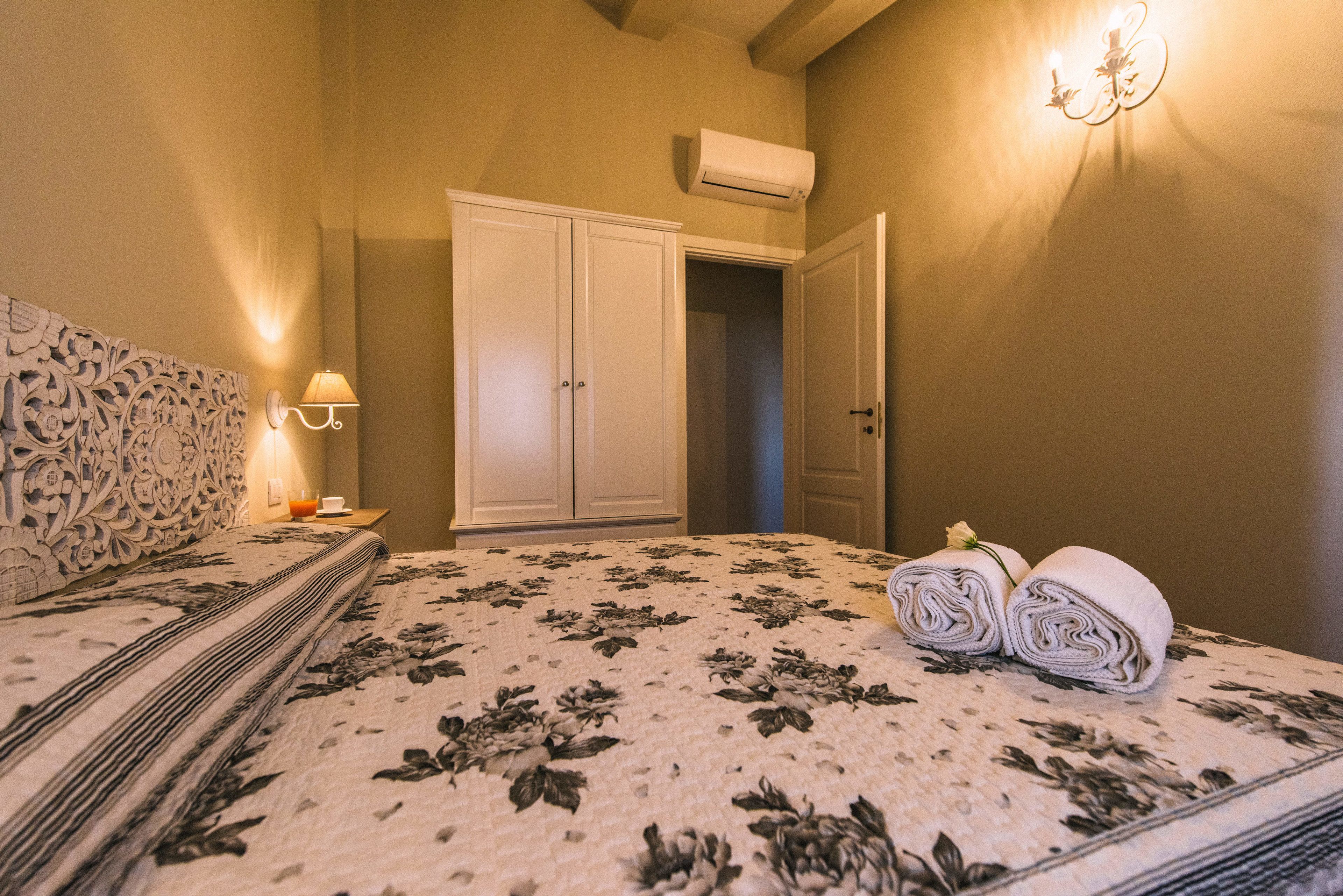 A bedroom with a floral bedspread, a bedside lamp, a rolled towel, and a wardrobe.