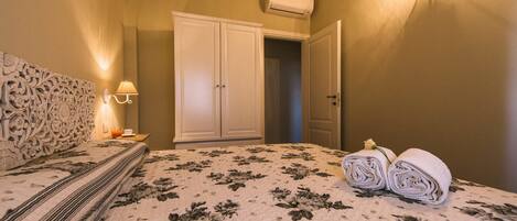 A bedroom with a floral bedspread, a bedside lamp, a rolled towel, and a wardrobe.
