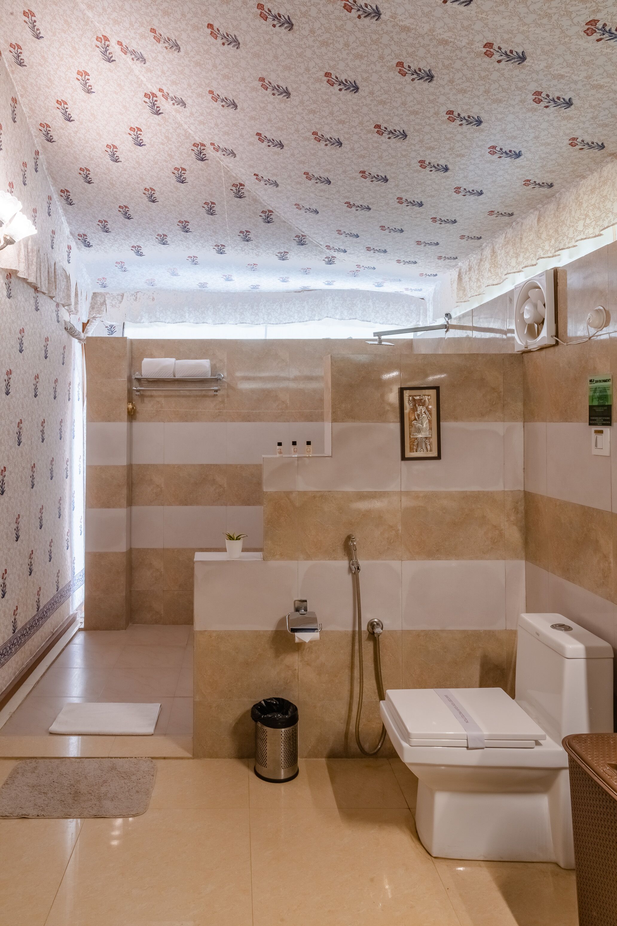 Une salle de bain avec douche, toilettes et lavabo, carrelée de carreaux beiges et blancs, avec un plafond à motifs et un petit tableau encadré accroché au mur.