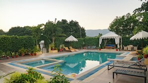 Piscina rectangular con borde de azulejos, rodeada de sillones y sombrillas, con un pabellón y plantas en macetas al fondo.