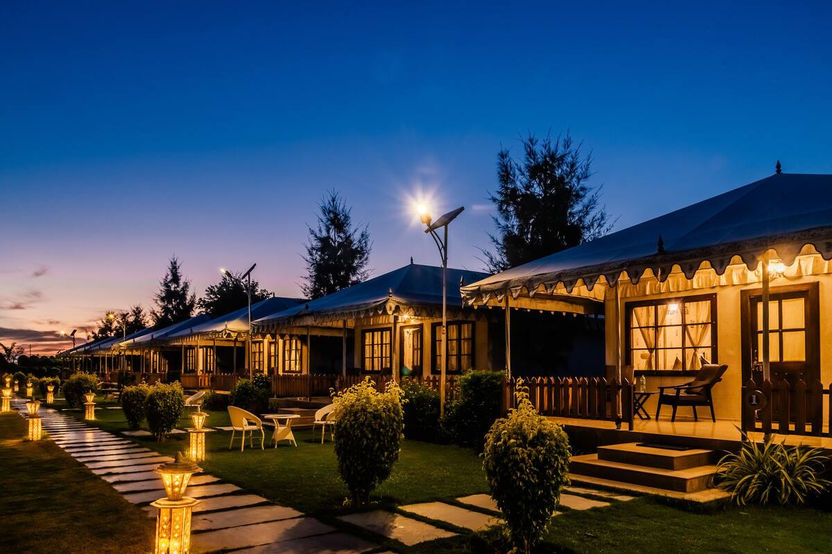 A row of cozy, well-lit cottages with outdoor seating areas, surrounded by neatly trimmed bushes and a stone pathway.