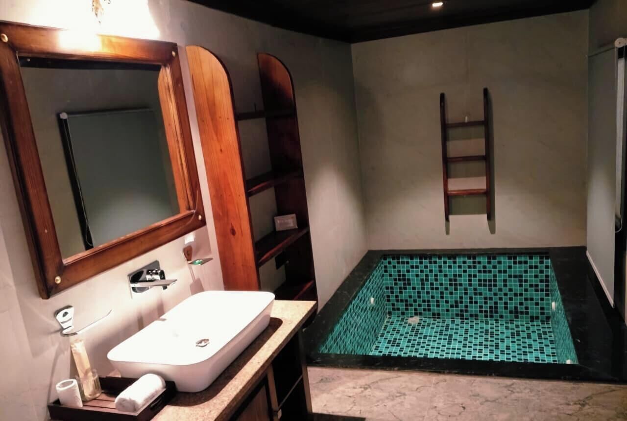 Une salle de bain avec un lavabo blanc, un miroir et une petite étagère. Une échelle est adossée au mur et le sol est carrelé.